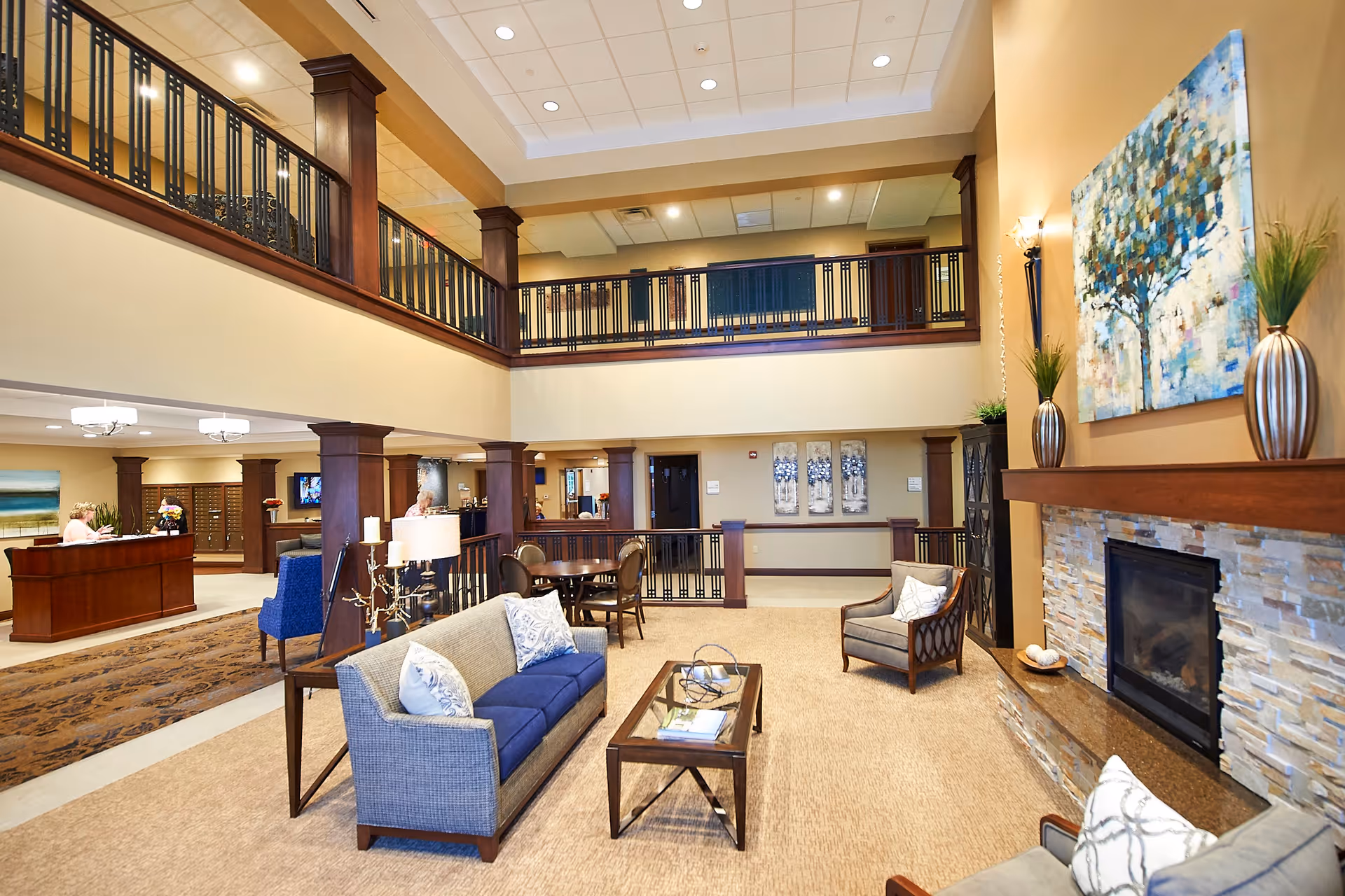 Spacious and well-lit senior living facility common area with a high ceiling and upper balcony. The room features comfortable seating including a blue sofa, armchairs, a coffee table, and a fireplace with a stone surround. There is a reception desk in the background and decorative artwork and plants enhancing the warm, inviting atmosphere.
