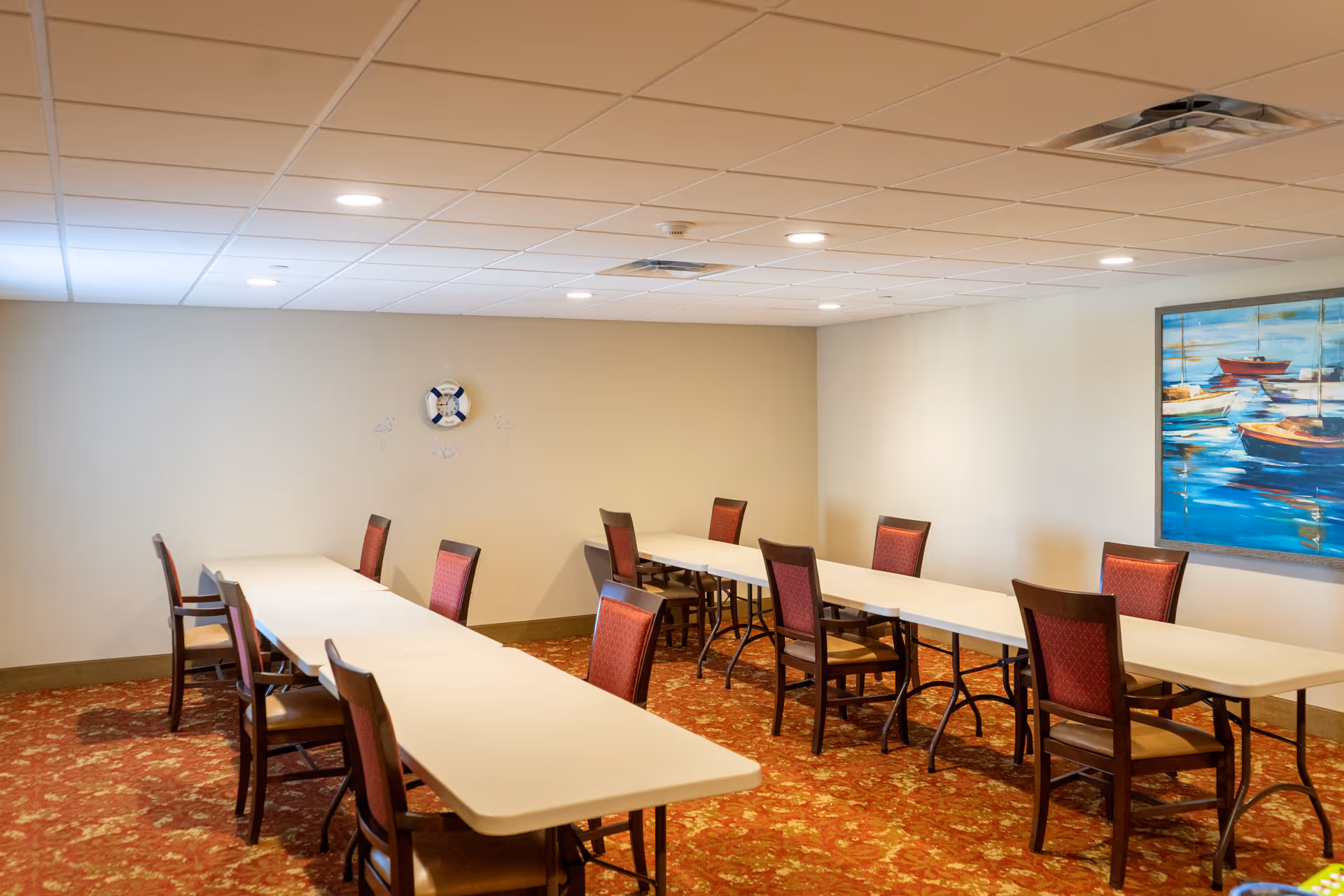 A meeting or activity room with two long white tables arranged in a U-shape, surrounded by wooden chairs with red cushioned seats and backs. The room has beige walls, a patterned red carpet, a ceiling with recessed lighting, a painting of boats on the water on one wall, and a decorative life preserver mounted on another wall.
