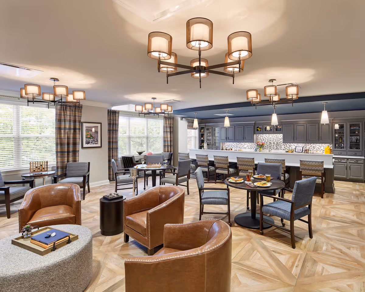 Open, well-lit dining and lounge area with tables, chairs, leather armchairs, and a long service counter/kitchen in the background.