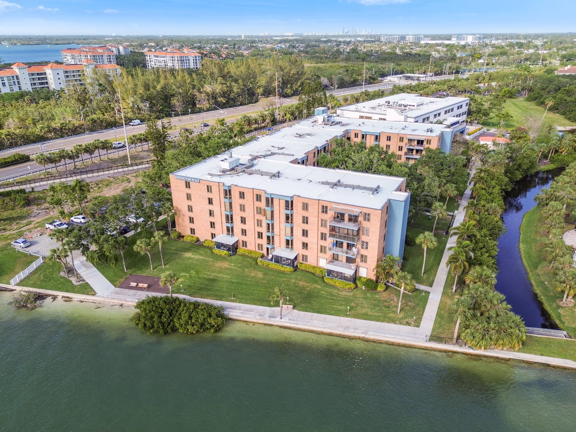 Aerial view of Addington Place at College Harbor, a multi-story residential building situated next to a body of water with a green lawn and trees surrounding it. There is a sidewalk along the water and a small canal on the right side. The background shows a road, more trees, and other buildings in the distance under a clear blue sky.