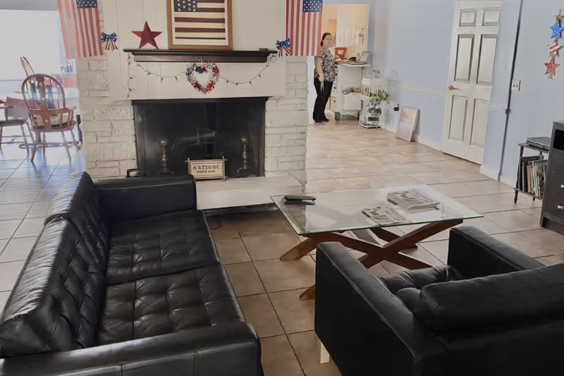 A living room area with black leather sofas and a glass coffee table in front of a white stone fireplace decorated with American flags and patriotic ornaments. A woman stands in the background near a hallway with light blue walls and tiled floors.