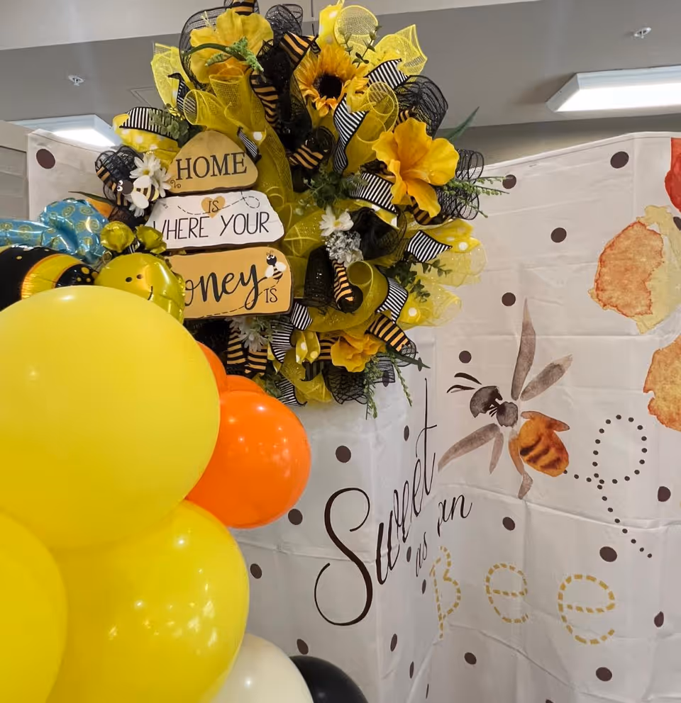 A decorative arrangement featuring a yellow and black wreath with flowers and ribbons, a sign that reads 'Home is where your honey is,' and a backdrop with a bee illustration and the phrase 'Sweet as can Bee.' There are also yellow, orange, white, and black balloons in the foreground.