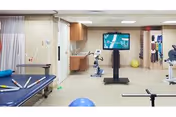 Physical therapy or rehabilitation room with exercise equipment including a stationary bike, therapy table with colorful rods, balance balls, and a large screen mounted on a stand displaying a virtual exercise or game. The room has beige walls, ceiling lights, and wooden cabinets.