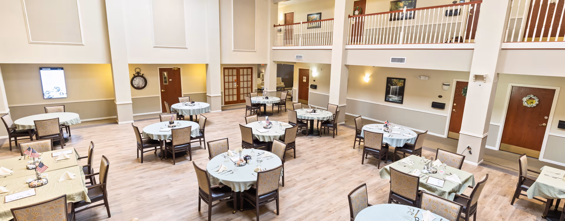 A spacious dining room with multiple round tables covered with light green tablecloths, each surrounded by chairs. The room has high ceilings with a second-floor balcony railing visible. The walls are painted in neutral tones with framed artwork and doors leading to other areas. The floor is light wood, and the tables are set with napkins, utensils, and small centerpieces.