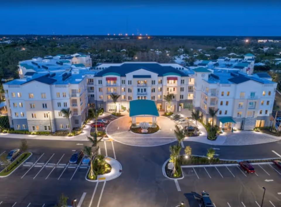 Aerial view of a large, well-lit senior living facility building at dusk with a circular driveway and parking lot in front, surrounded by trees and residential areas.