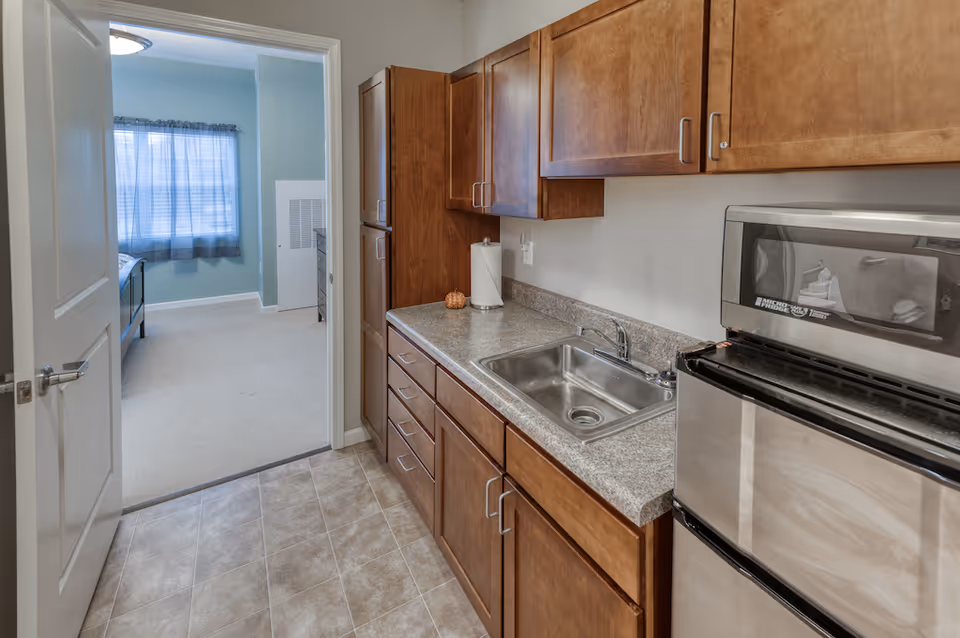 View of a small kitchen area with wooden cabinets, a stainless steel sink, and a compact refrigerator and microwave. The kitchen floor is tiled, and through an open door, a bedroom with a bed and a window with blue curtains is visible.