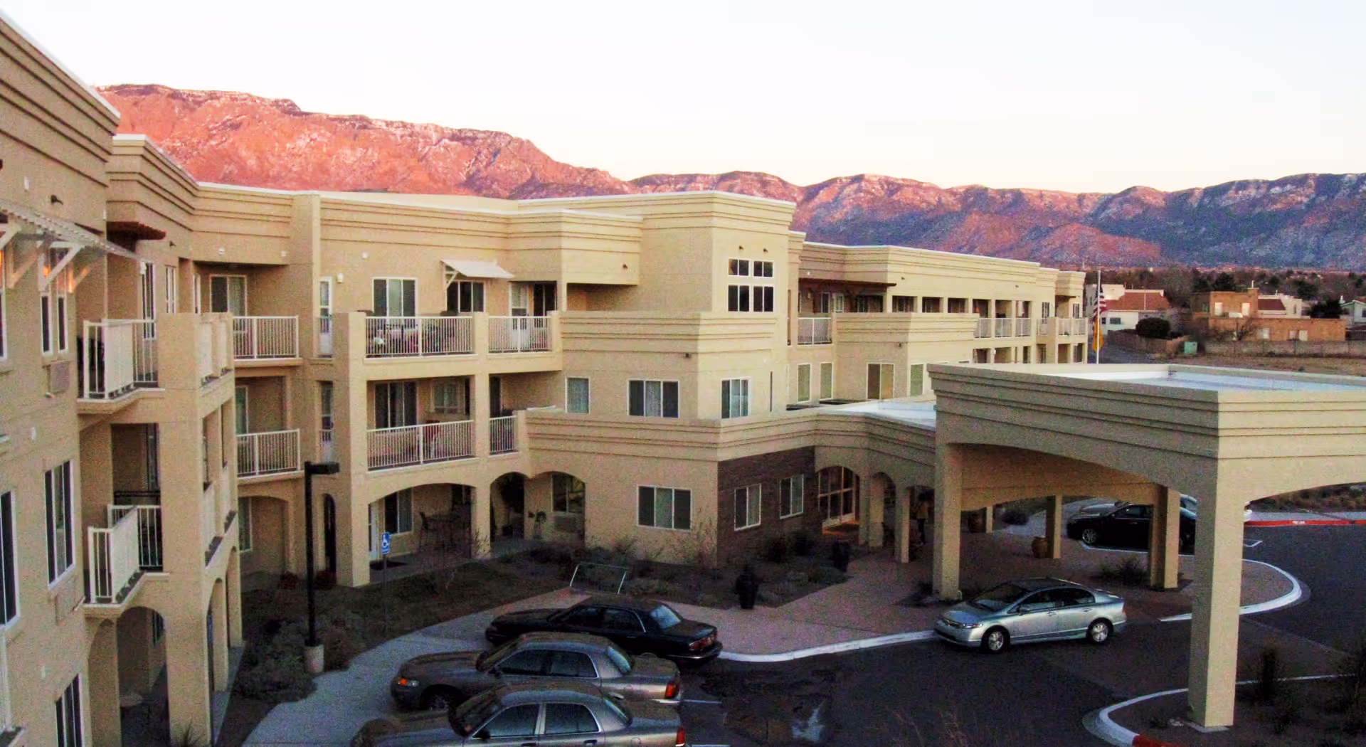 Exterior view of a multi-story beige retirement living facility with balconies, a covered entrance, parked cars, and mountains in the background during sunset.