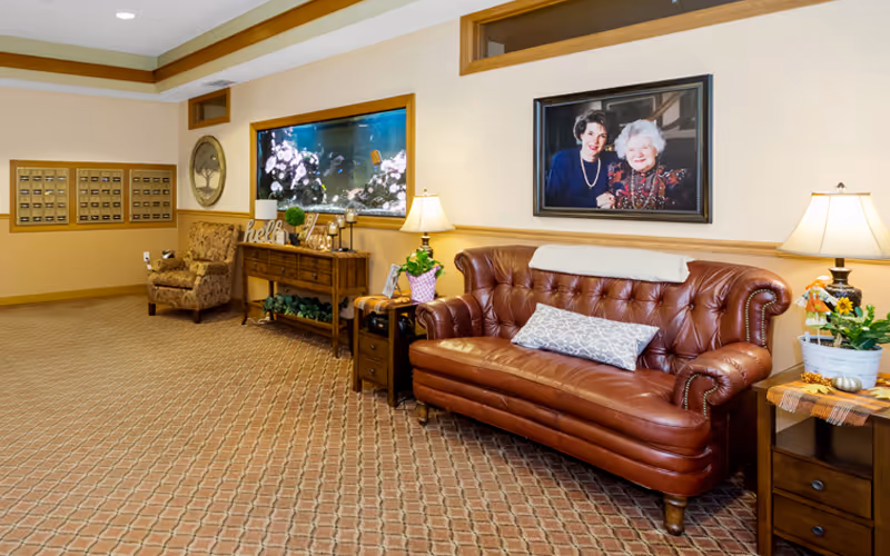 Cozy reception lounge with a brown leather sofa, armchair, side tables with lamps, a large wall aquarium and mailboxes along a carpeted hallway.