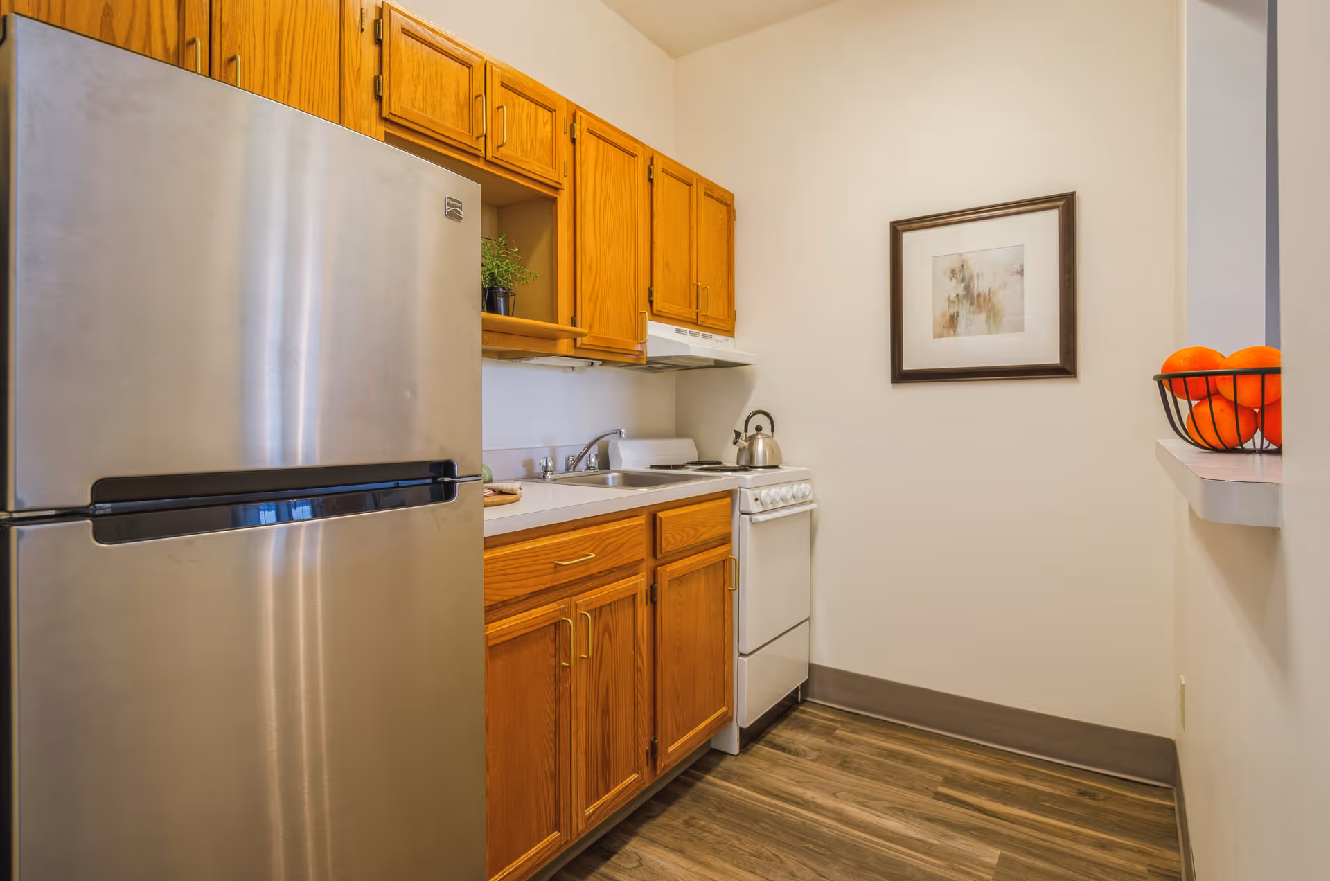 A small kitchen with wooden cabinets, a stainless steel refrigerator, a white stove with a kettle on it, a sink, and a basket of oranges on a counter. There is a framed abstract painting on the wall.