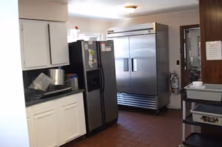 Interior view of a kitchen area with white cabinets, a black refrigerator, a large stainless steel commercial refrigerator, and a metal cart with dishes on it. The floor is covered with reddish-brown tiles.