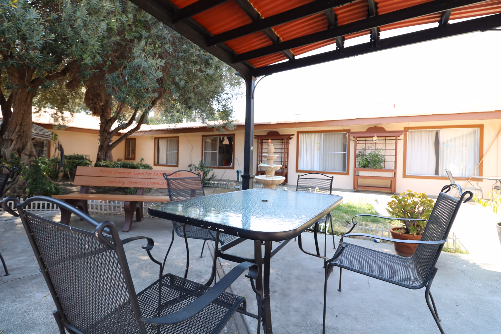 Outdoor seating area at Willow Pass Healthcare Center featuring a glass-top metal table with four metal chairs under a red-roofed pergola. In the background, there is a beige building with windows and a small fountain surrounded by greenery and potted plants.