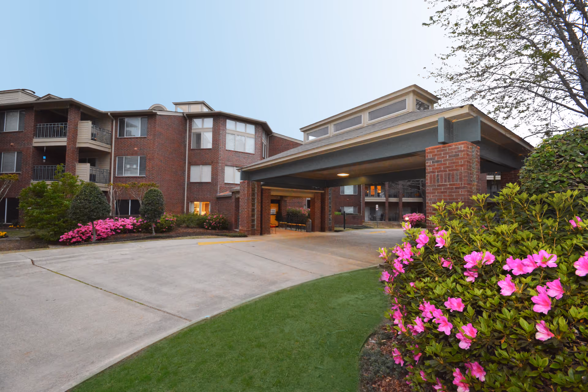 Exterior view of a multi-story brick building with a covered entrance driveway. The building is surrounded by well-maintained landscaping including green bushes and vibrant pink flowers. The sky is clear and the scene appears to be taken during daylight.