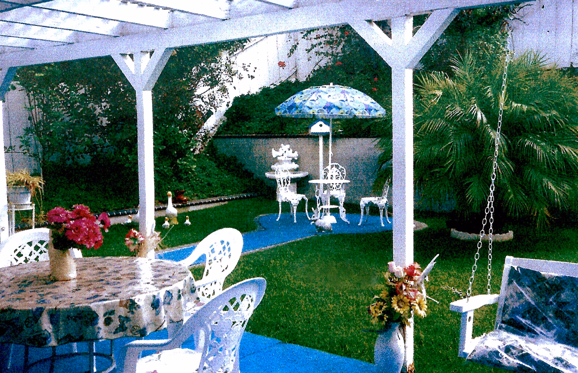 A covered outdoor patio area with a round table covered by a floral tablecloth and surrounded by white plastic chairs. There are flower vases on the table and nearby. Beyond the patio is a small garden with green grass, plants, and a white metal table with chairs under a blue patterned umbrella. A white swing chair is partially visible on the right side.