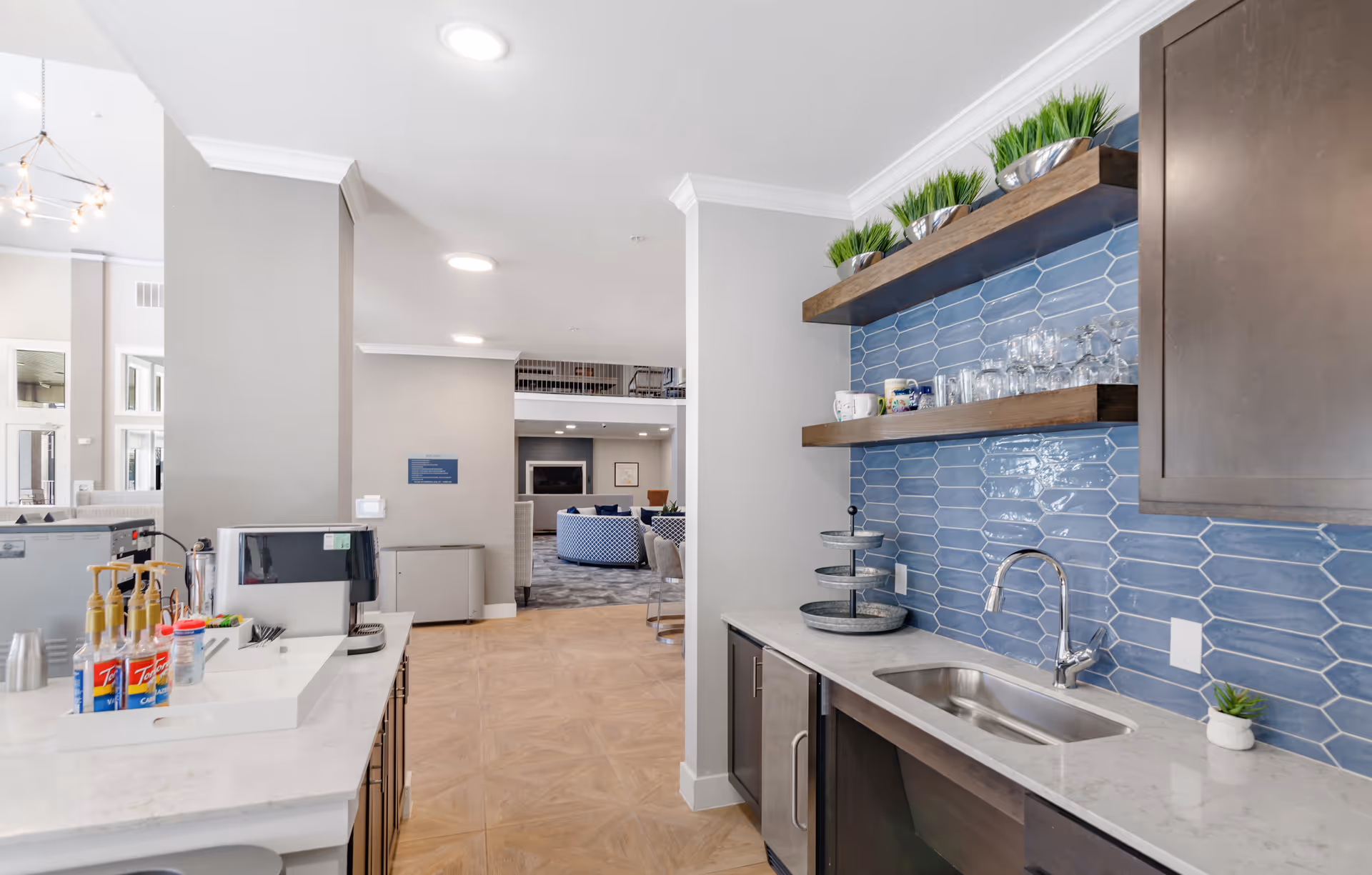 Interior view of a modern kitchenette area with a marble countertop, stainless steel sink, blue hexagonal tile backsplash, and wooden shelves holding glassware and small plants. To the left, there is a coffee machine and syrup bottles on the counter. In the background, a lounge area with blue and white patterned chairs and a fireplace is visible.