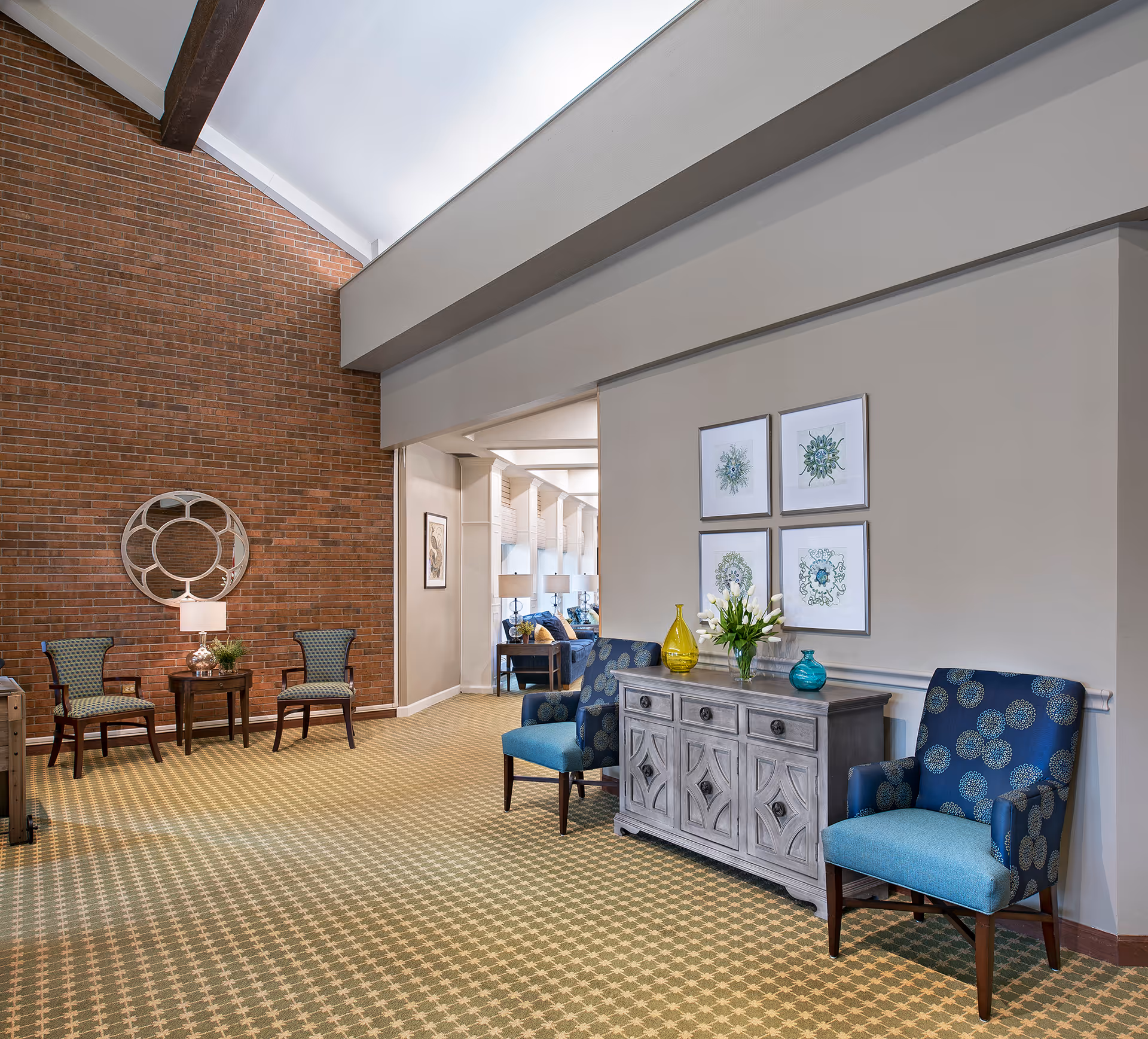 Bright senior living lounge with patterned chairs, a decorative console table, and a brick accent wall.