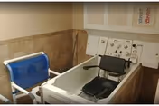 Accessible bathing area with a walk-in tub containing a transfer chair, an adjacent blue padded bath lift, tiled walls and wall-mounted control panels.