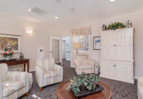 A cozy living room with three striped armchairs arranged around a round wooden coffee table with a green plant centerpiece. The room features a white cabinet with decorative items on top, a side table with a lamp, framed artwork on the walls, and a door with glass panels leading to another room.