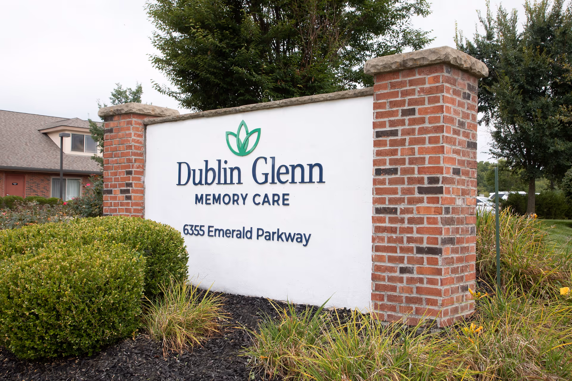 A large white sign with brick pillars on each side displaying the name 'Dublin Glenn Memory Care' and the address '6355 Emerald Parkway', surrounded by green bushes and plants with a building and trees in the background.