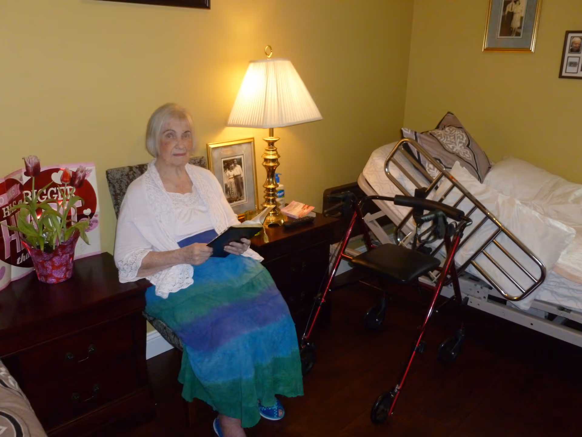 An elderly woman sitting on a chair in a bedroom, holding a book. The room has a bedside table with a lamp, framed photos, a flower pot, and a walker in front of an adjustable bed with pillows.