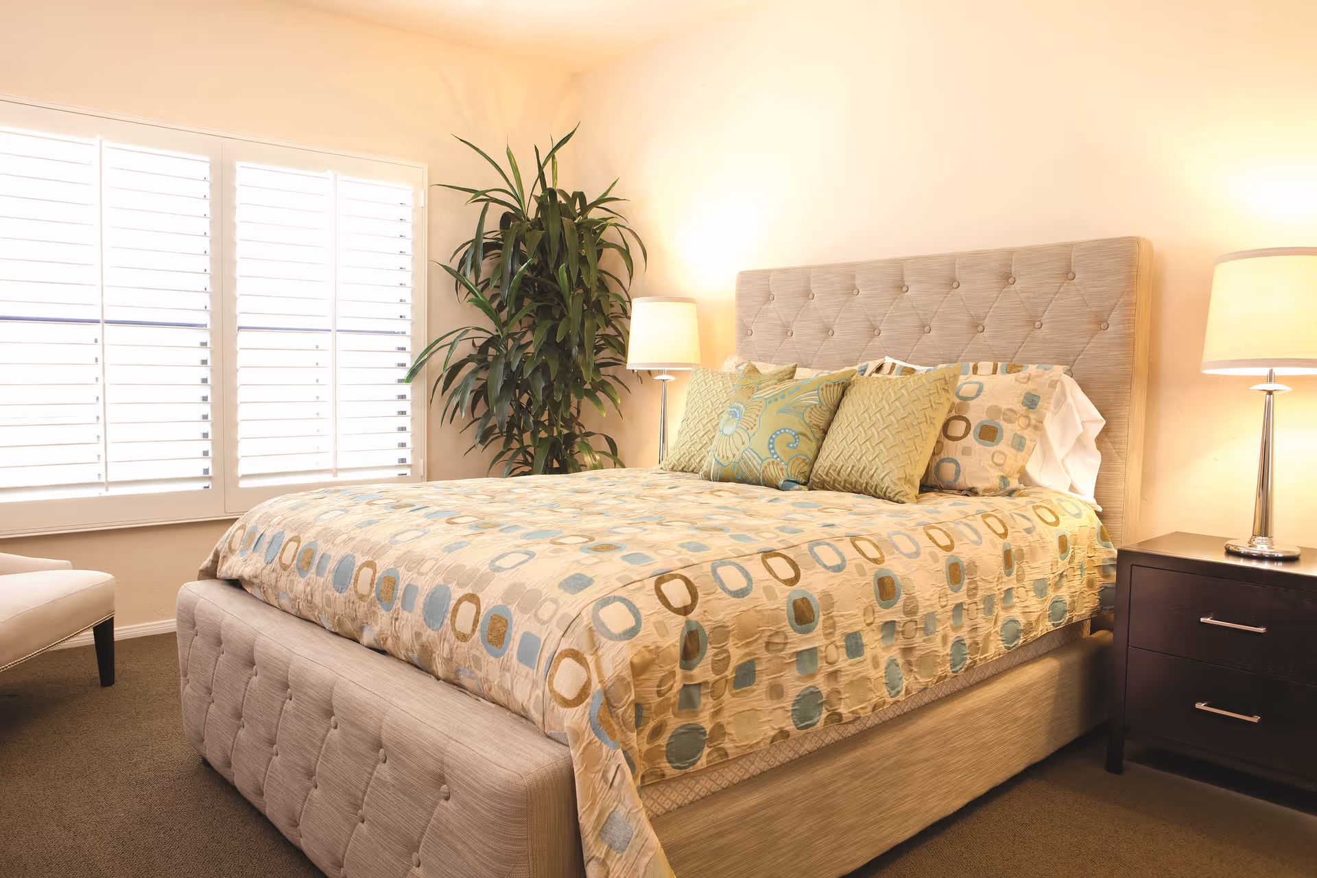 A well-lit bedroom featuring a large bed with a tufted beige headboard and matching footboard. The bed is covered with a patterned comforter in shades of beige, blue, and brown, and is adorned with several decorative pillows. On the right side of the bed is a dark wooden nightstand with two drawers and a modern lamp with a beige shade. A large green potted plant is positioned in the corner near a window with white plantation shutters. The room has beige walls and carpeted flooring.