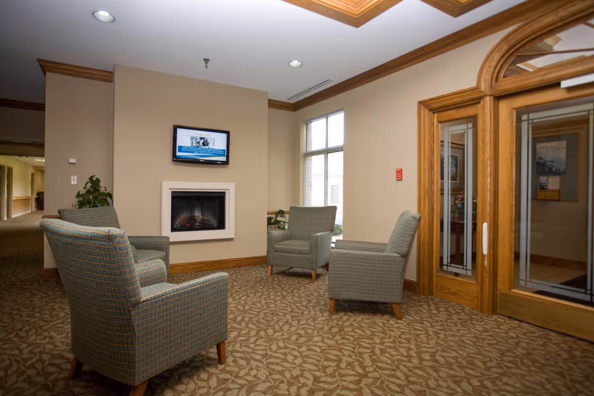 A cozy seating area in a healthcare facility with four upholstered armchairs arranged around a wall-mounted electric fireplace. Above the fireplace is a flat-screen TV. The room has beige walls, a patterned carpet, large windows letting in natural light, and wooden framed glass doors on the right.