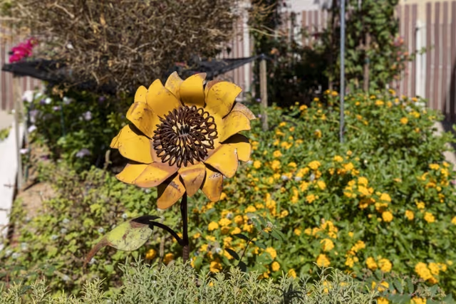 A close-up of a decorative yellow metal sunflower in a garden with green plants and yellow flowers in the background, along with a wooden fence and some other plants.