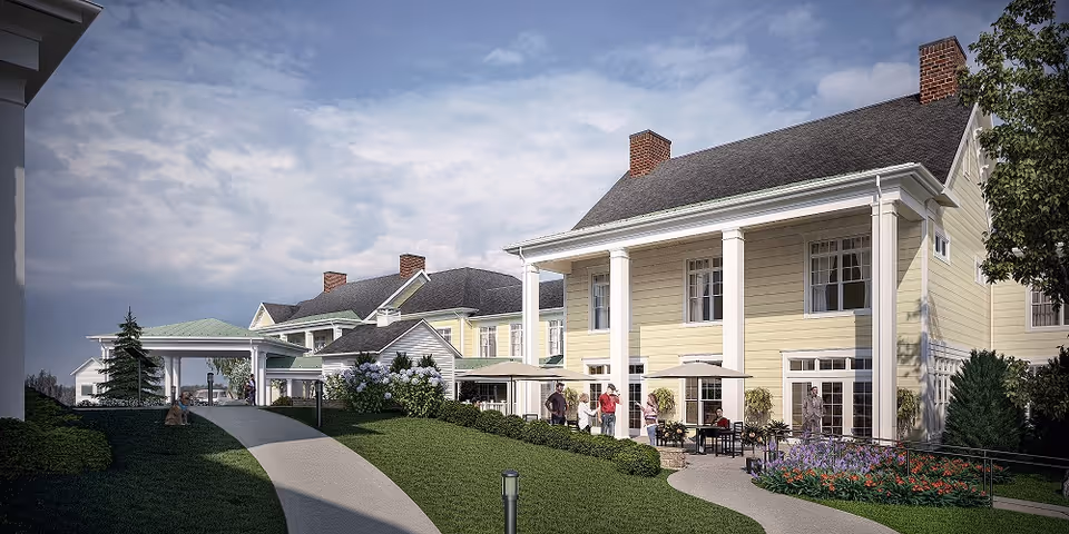 Exterior view of a large, yellow senior living facility building with white columns and multiple windows. There is a paved walkway leading up to the entrance, surrounded by green grass, bushes, and colorful flowers. Several people are standing and sitting under umbrellas on the patio area. The sky is partly cloudy.