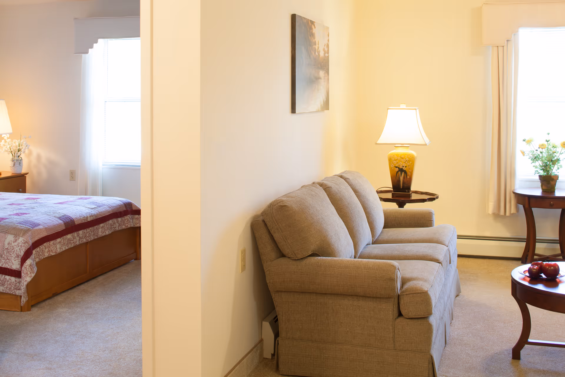 A cozy living area with a beige sofa, a side table with a yellow lamp, and a round wooden coffee table with red apples. Adjacent to the living area is a bedroom with a bed covered in a patterned quilt, a wooden nightstand with a lamp, and a window with white curtains.