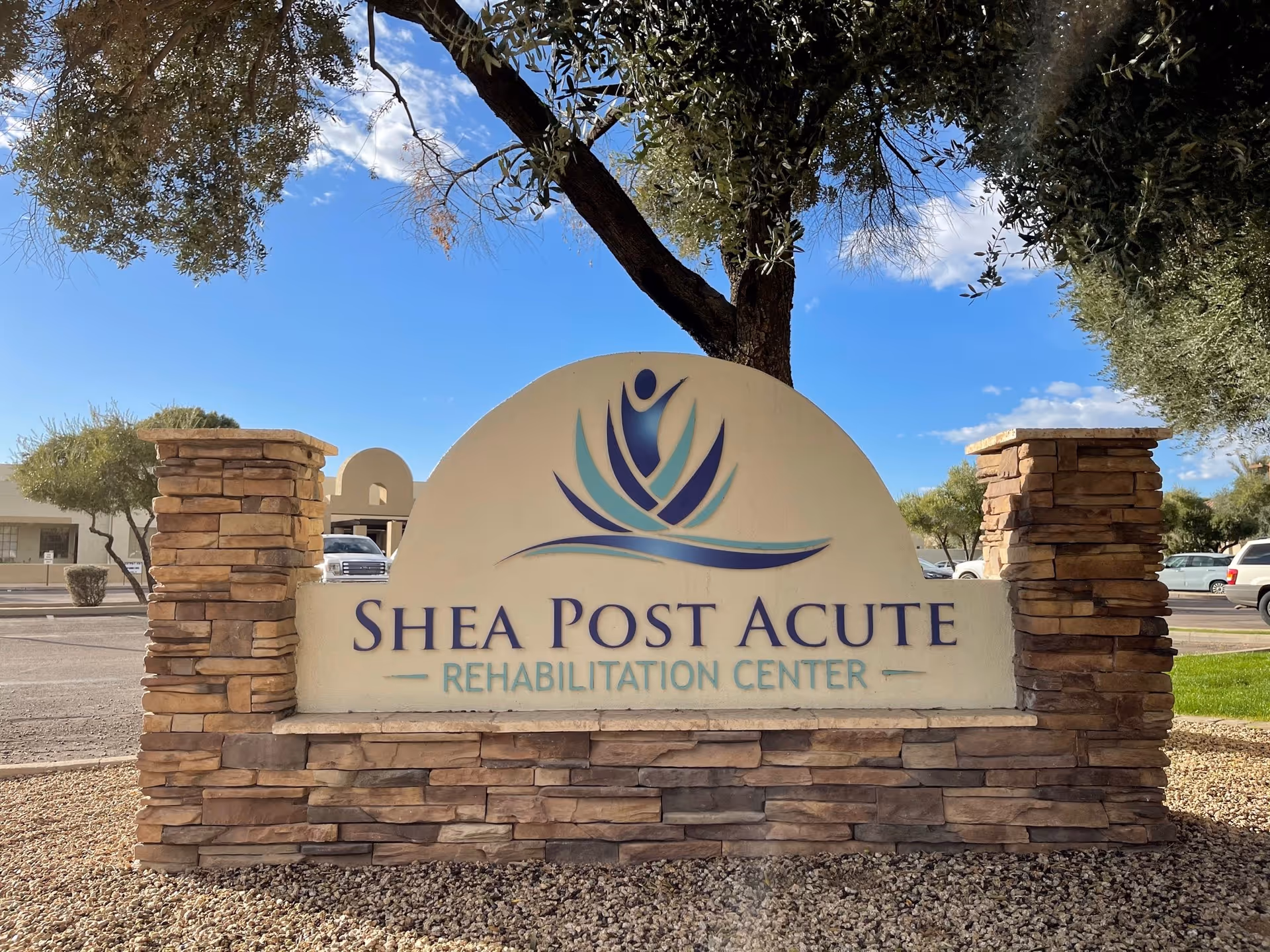 A stone entrance sign that reads "Shea Post Acute Rehabilitation Center" with trees, parked cars, and a blue sky in the background.