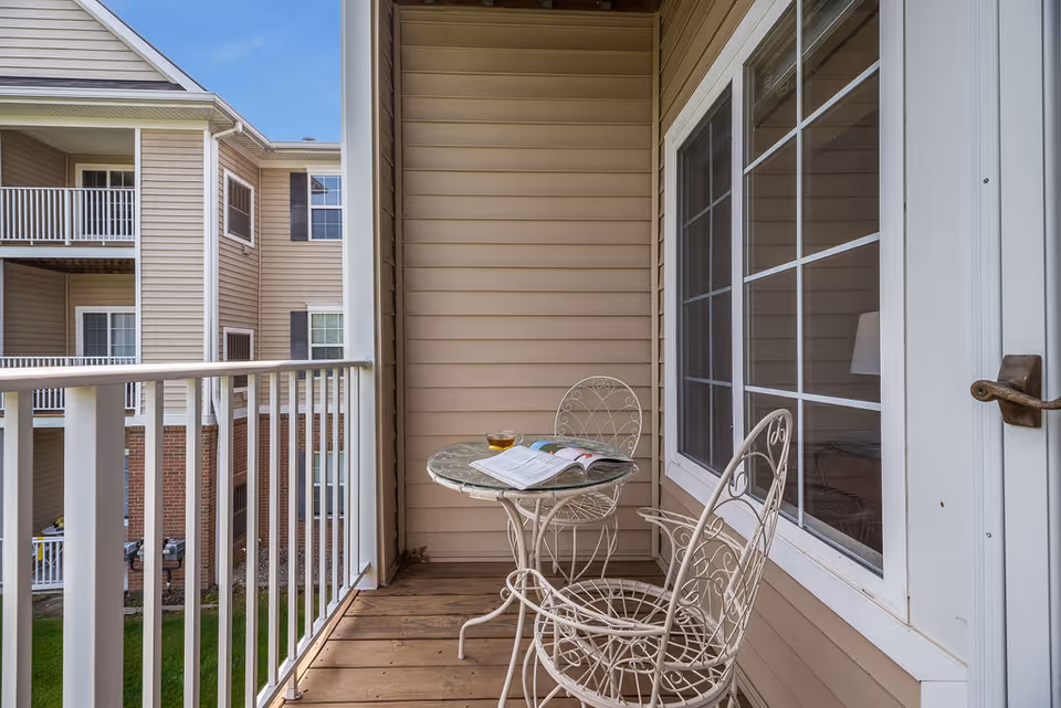 Balcony with white wrought-iron table and two chairs, an open book and a drink, overlooking adjacent apartment balconies.