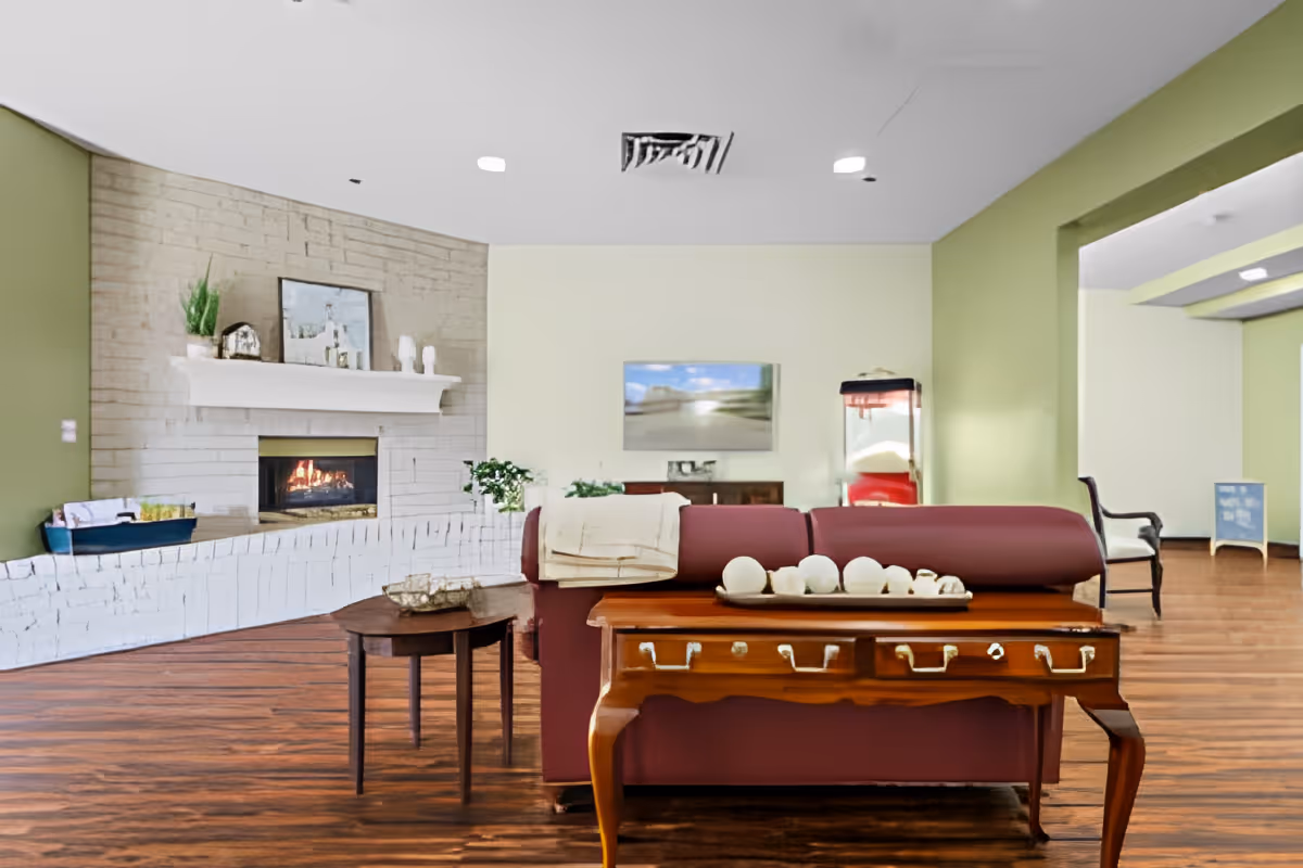 A cozy living room area with a lit fireplace set in a white brick wall. There is a maroon sofa with a wooden console table behind it, decorated with white spherical objects. A small round side table is next to the sofa. The walls are painted light green and cream, and there is a popcorn machine in the corner. The floor is wooden, and there is a TV mounted on the cream wall.