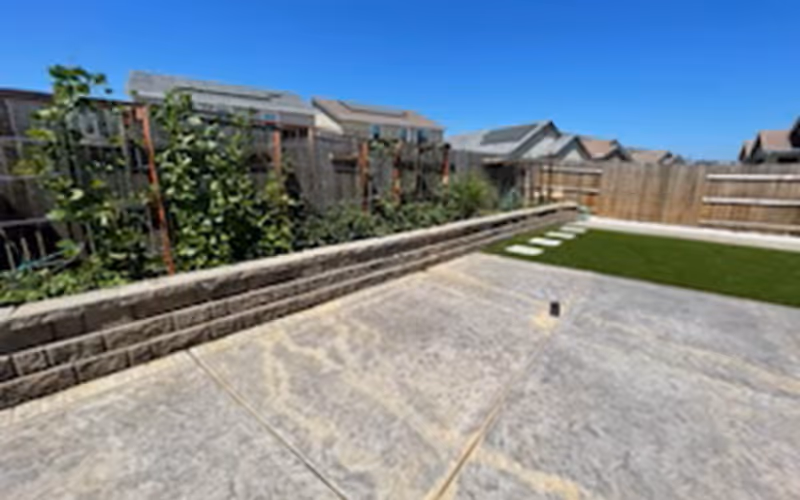 Sunlit fenced backyard featuring a stamped concrete patio, a raised stone planter with plants, and a small artificial turf lawn with stepping stones.