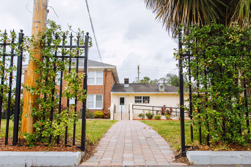 View through an open black metal gate with green climbing plants on both sides, leading to a paved walkway towards a two-story brick and white building under a cloudy sky.