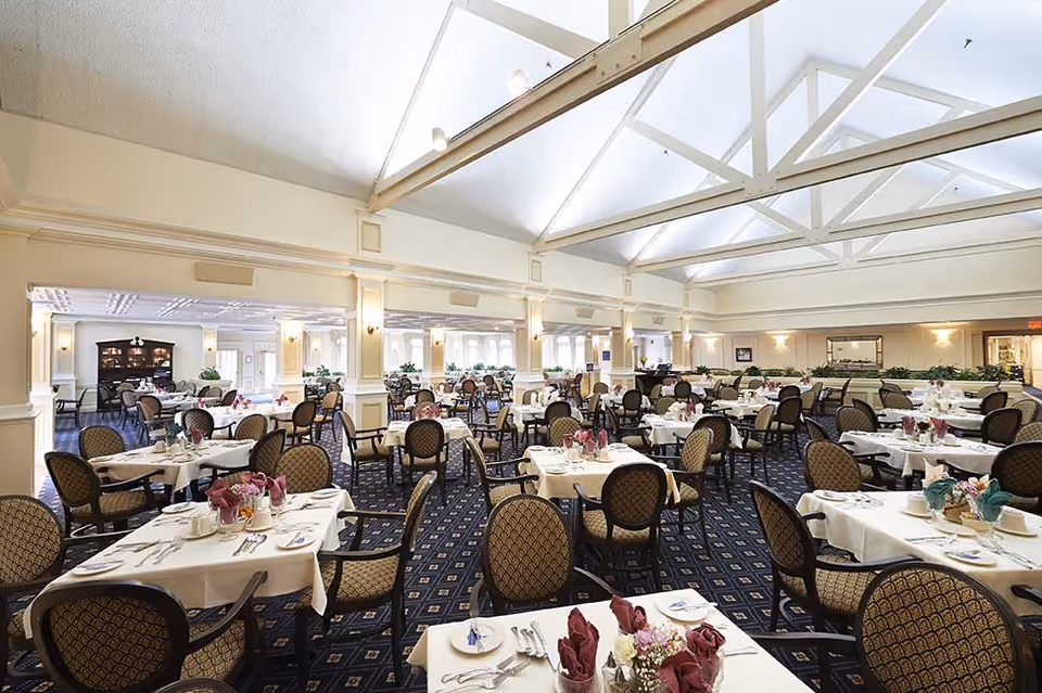 Spacious dining room with multiple tables covered in white tablecloths, each set with plates, silverware, and folded napkins. The room features high ceilings with exposed beams and large windows allowing natural light. Upholstered chairs with patterned fabric surround the tables, and there are plants and decorative elements along the walls.