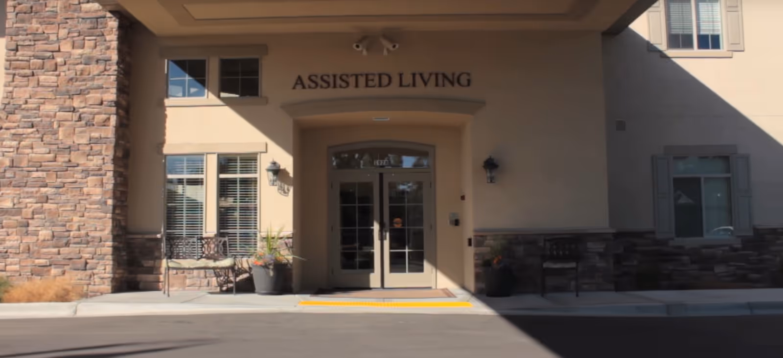 Entrance to an assisted living facility with double glass doors under a covered entryway. The building exterior features stone and beige stucco walls, two benches, potted plants, and windows with shutters. The words 'ASSISTED LIVING' are displayed above the door.