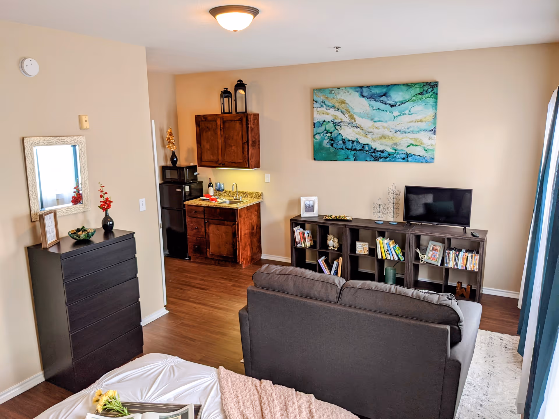 A cozy studio apartment interior featuring a bed with a pink throw blanket and flowers, a dark gray sofa facing a TV on a bookshelf filled with books and decorative items, a small kitchenette with wooden cabinets, a granite countertop, a microwave, and a mini fridge. The walls are beige with a large abstract painting above the TV, and a window with blue curtains lets in natural light.