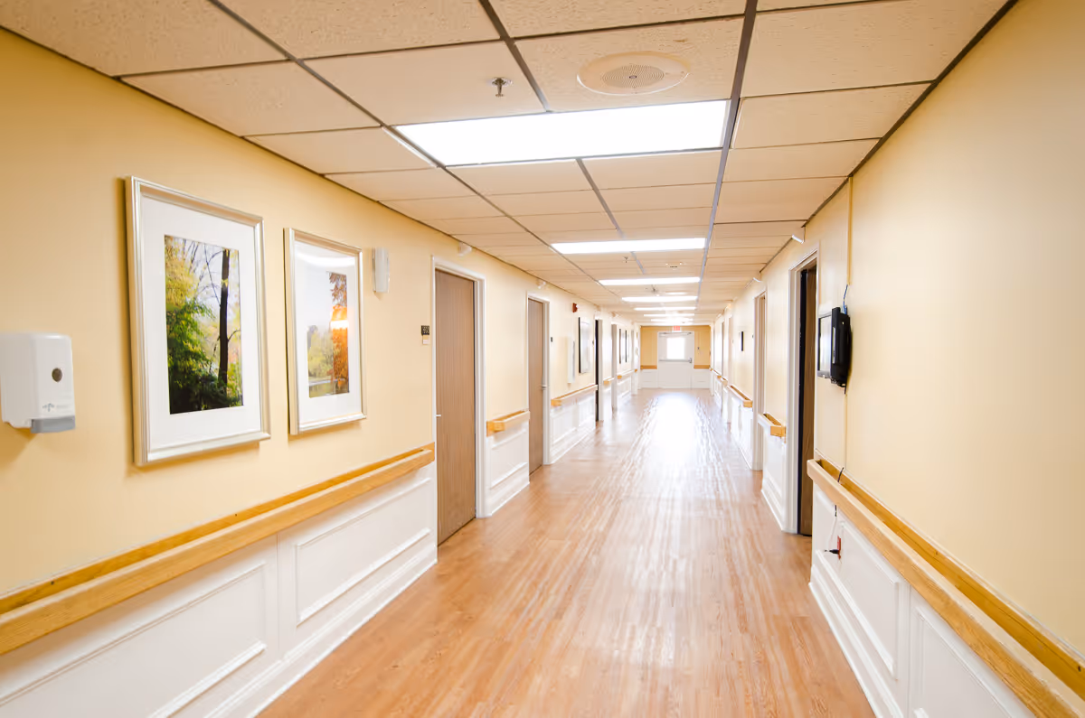 A long, well-lit hallway in a senior living facility with light yellow walls, wooden handrails on both sides, framed nature photographs on the left wall, and multiple closed doors along the corridor. The floor is wood-patterned, and the ceiling has recessed lighting panels.