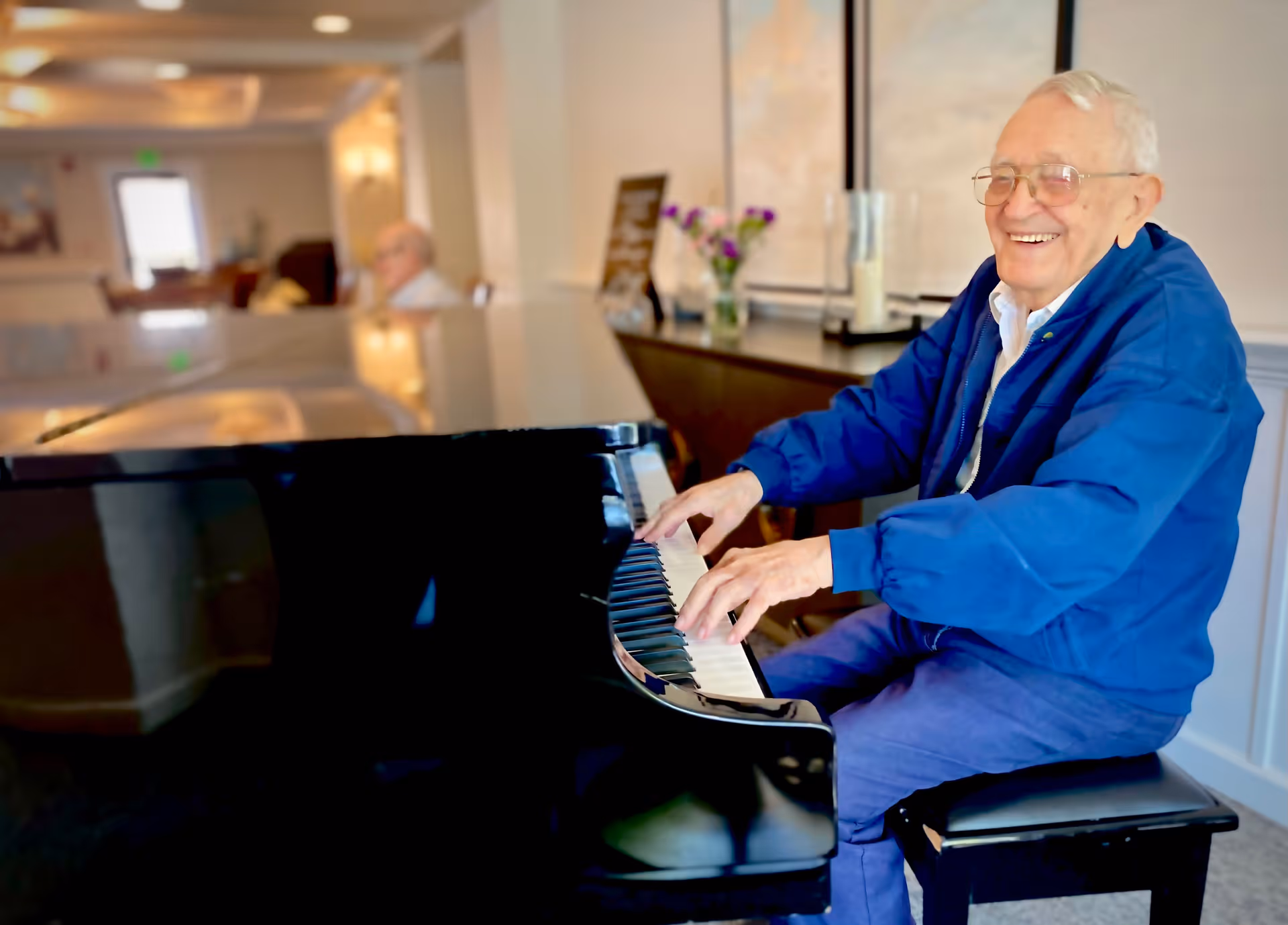 An elderly man wearing glasses and a blue jacket is smiling while playing a black grand piano in a well-lit room with soft lighting and decorative elements in the background.