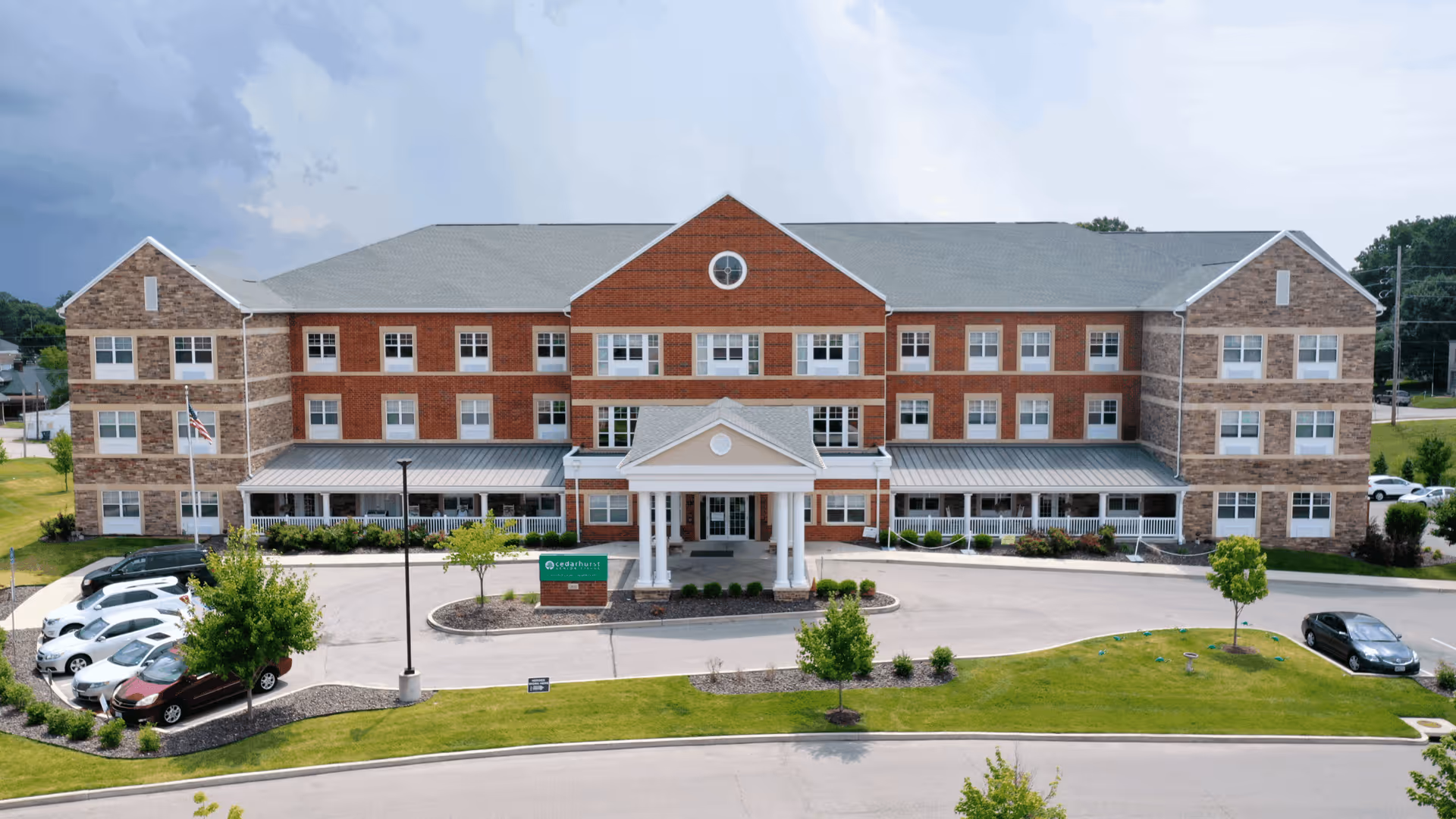 Front exterior view of Cedarhurst Senior Living of St. Charles, a three-story brick building with a covered entrance supported by white columns, surrounded by a parking lot with several cars and landscaped greenery.