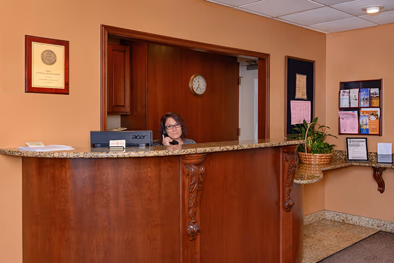 Reception area with a wooden front desk and granite countertop. A woman is sitting behind the desk, talking on the phone. The wall behind her has a clock and a doorway leading to another room. On the right side, there is a bulletin board with various notices and a basket with a plant on a small counter.