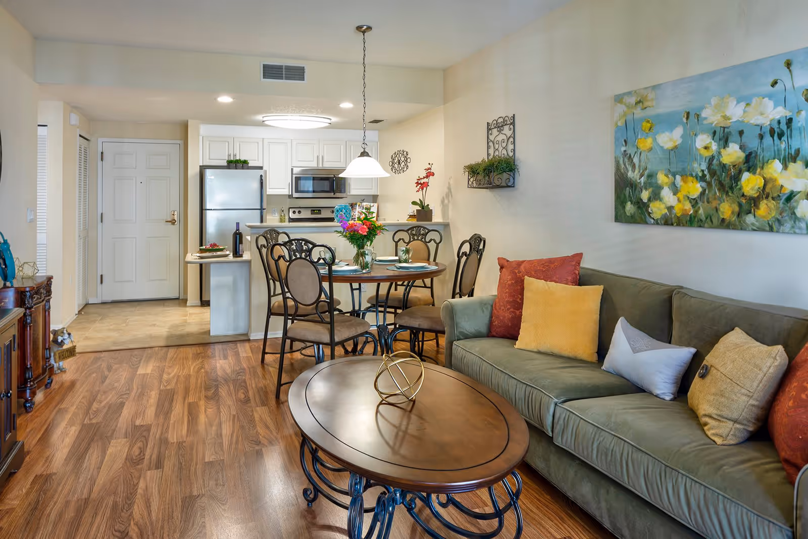 A cozy living room and kitchen area in an apartment. The living room features a green sofa with colorful pillows and a wooden coffee table with a decorative item. The dining area has a round table with four chairs and a vase of flowers. The kitchen includes white cabinets, a refrigerator, microwave, and stove. The walls are decorated with a floral painting and a small wall-mounted plant holder.