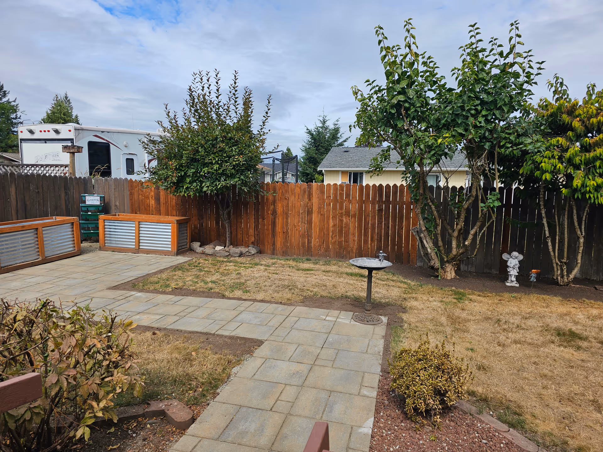 A backyard area with a paved walkway, dry grass patches, a birdbath, several trees, and a wooden fence. There are two raised garden beds with wooden and metal sides, and a small garden statue near the fence. A white RV is visible behind the fence on the left side.