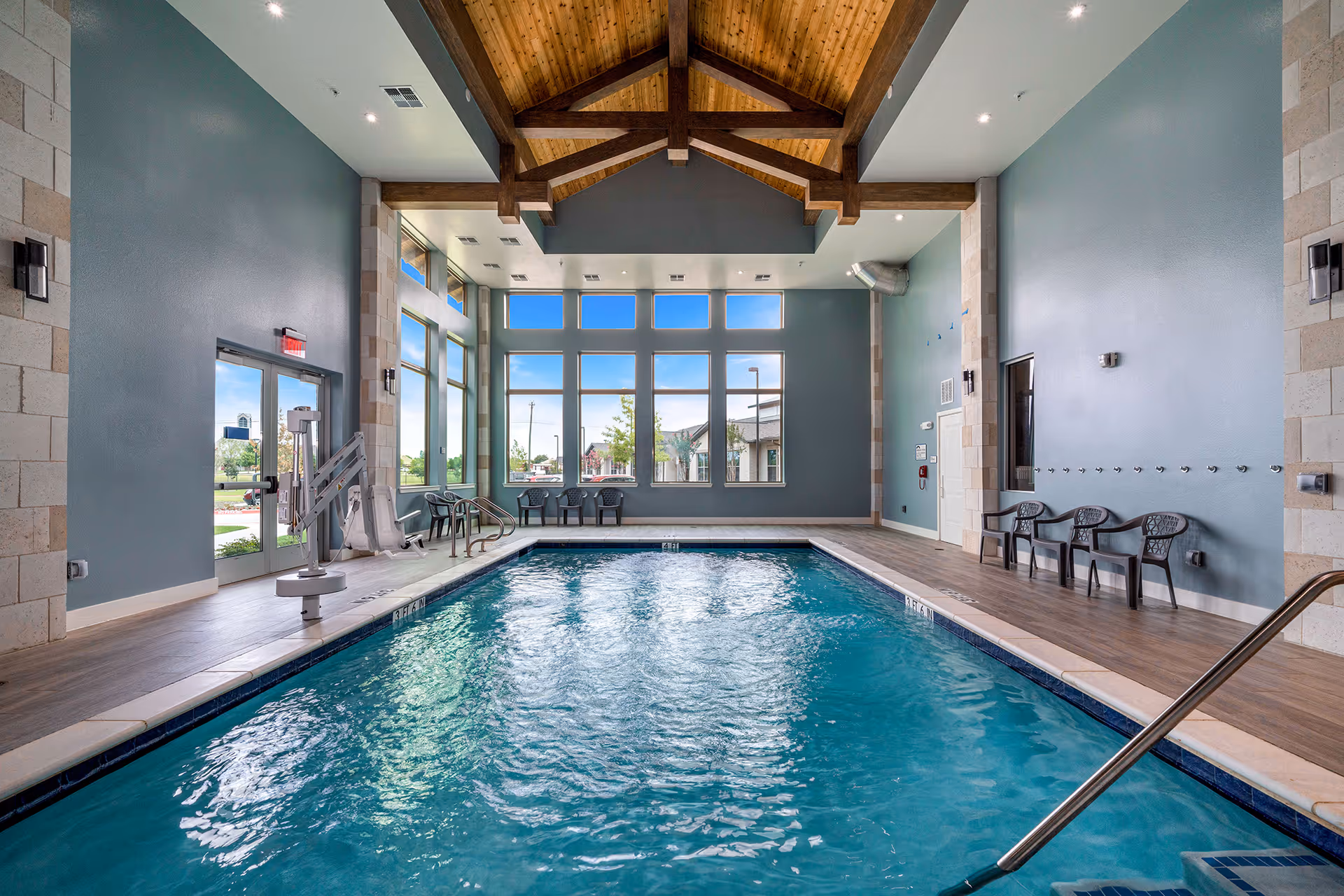 Indoor swimming pool area with clear blue water, surrounded by a wooden floor and teal walls. The ceiling features exposed wooden beams and large windows letting in natural light. Several plastic chairs are lined up along the walls, and there is a pool lift for accessibility near the entrance door.