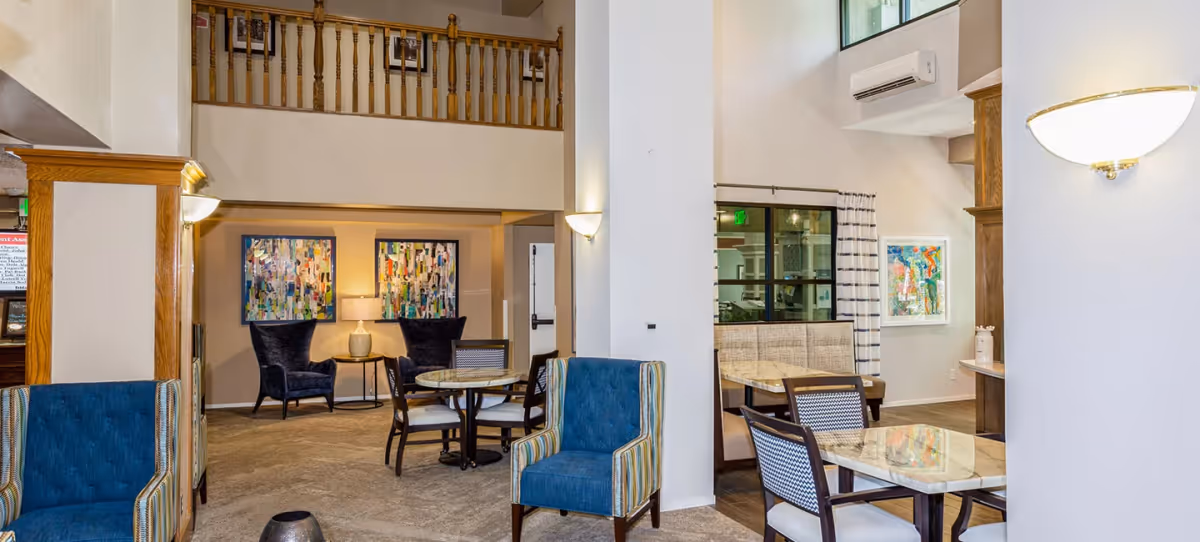 A spacious and well-lit common area in The Windham senior living facility featuring comfortable blue and striped armchairs, round tables with chairs, a booth seating area, colorful abstract artwork on the walls, and warm lighting fixtures. The space has a high ceiling with a wooden railing on the upper level and a split air conditioning unit mounted on the wall.