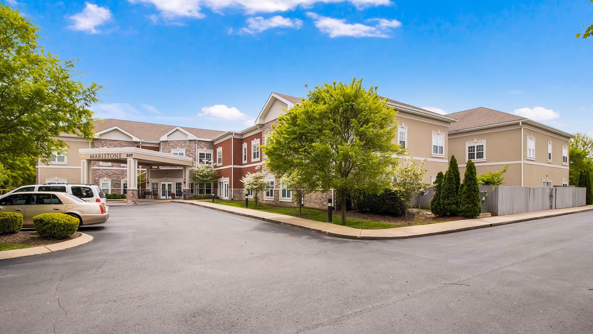 Front exterior of the Maristone of Franklin senior living building with an entrance canopy, parked cars, and landscaped trees.