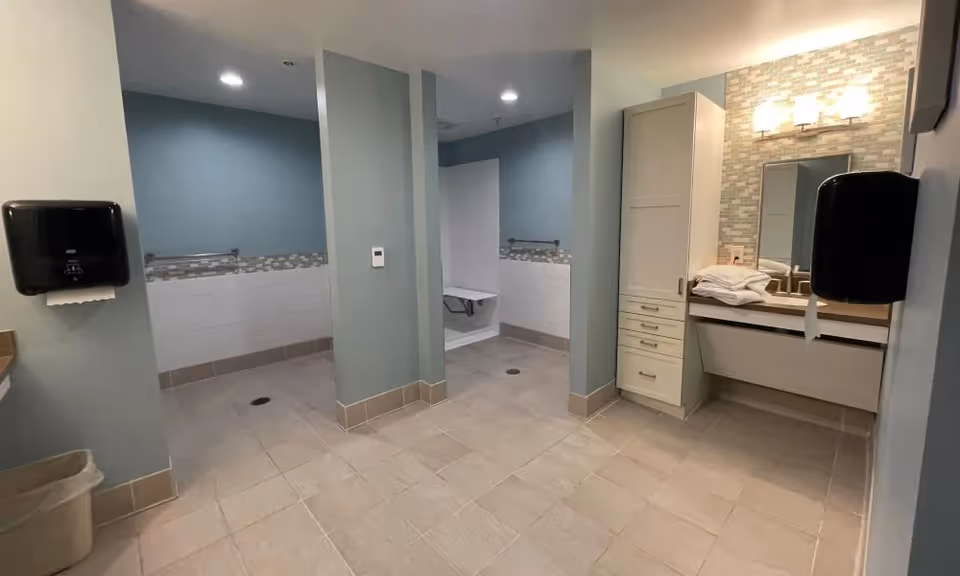A spacious, modern bathroom with light blue walls and beige tiled floors. The room features two open shower areas with grab bars and a fold-down seat in one shower. There is a vanity with a large mirror, cabinet storage, and neatly folded towels. Two black paper towel dispensers are mounted on the walls, and a trash can is visible in the corner.