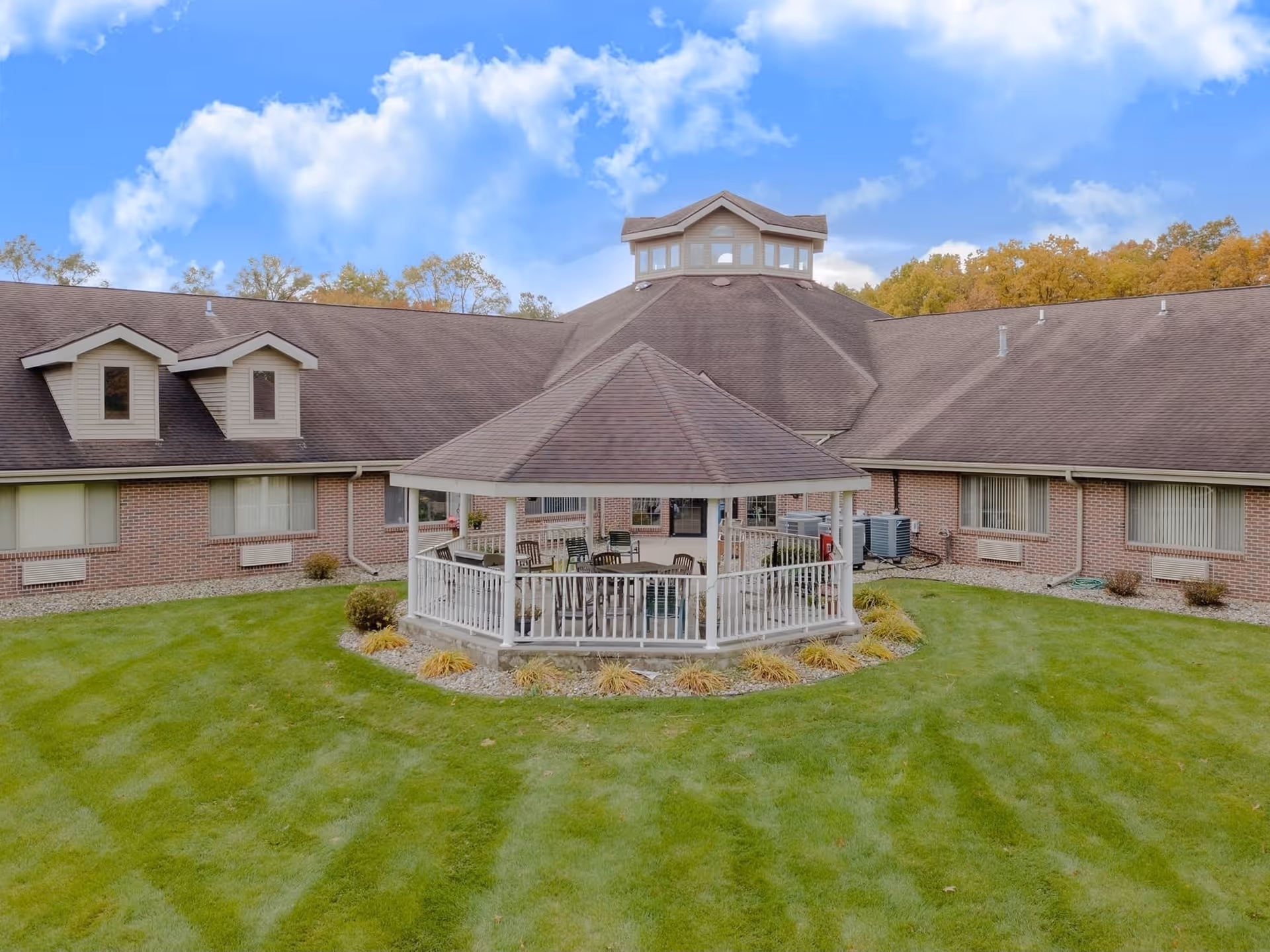 Outdoor view of a senior living facility with a large green lawn and a white gazebo in the center. The building is made of brick with a brown roof and multiple windows. Trees with autumn foliage are visible in the background under a partly cloudy blue sky.