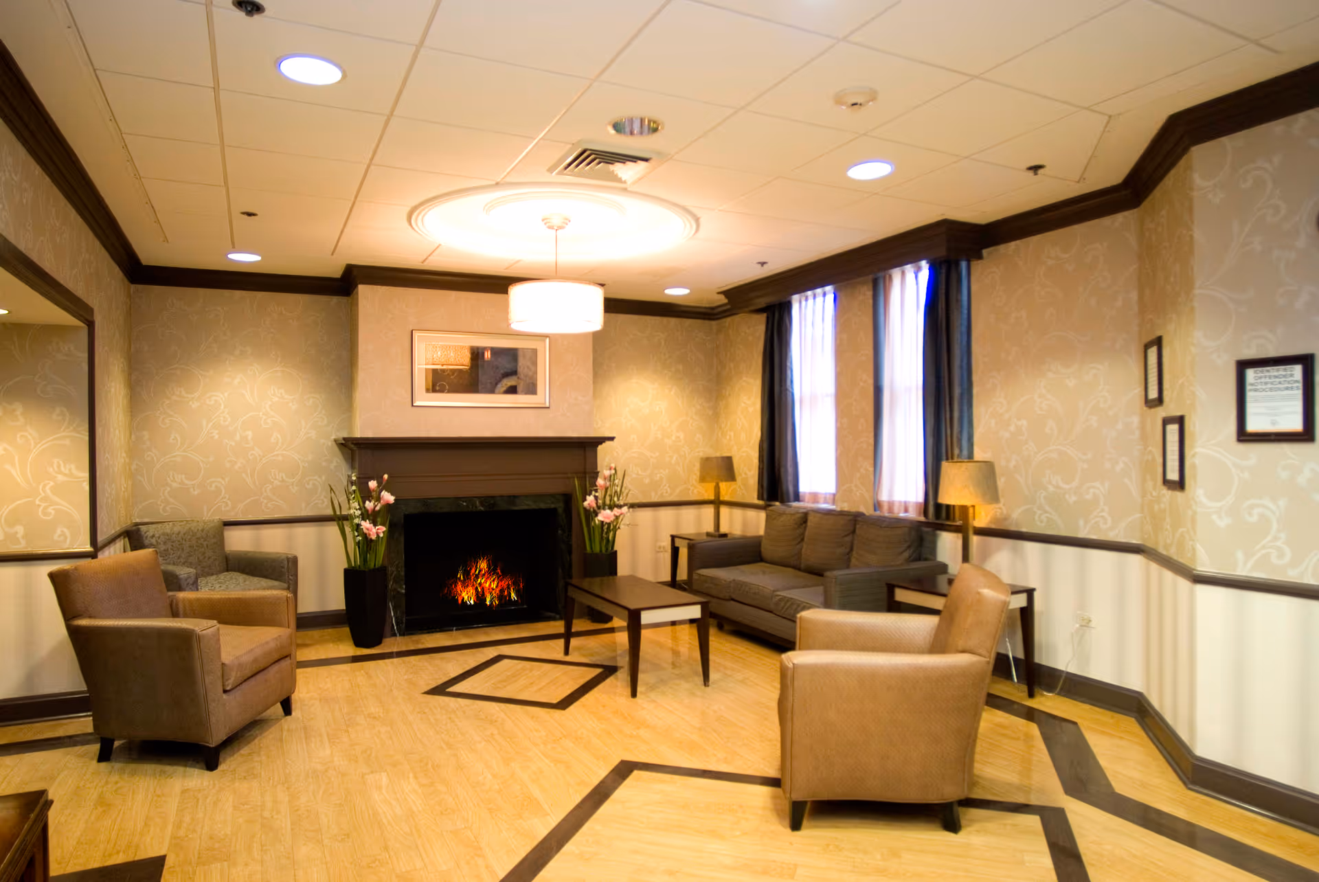 A cozy living room with a fireplace, two armchairs, a sofa, a coffee table, two side tables with lamps, and two tall flower vases on either side of the fireplace. The room has patterned wallpaper, wooden flooring with dark inlays, and two windows with curtains letting in natural light.
