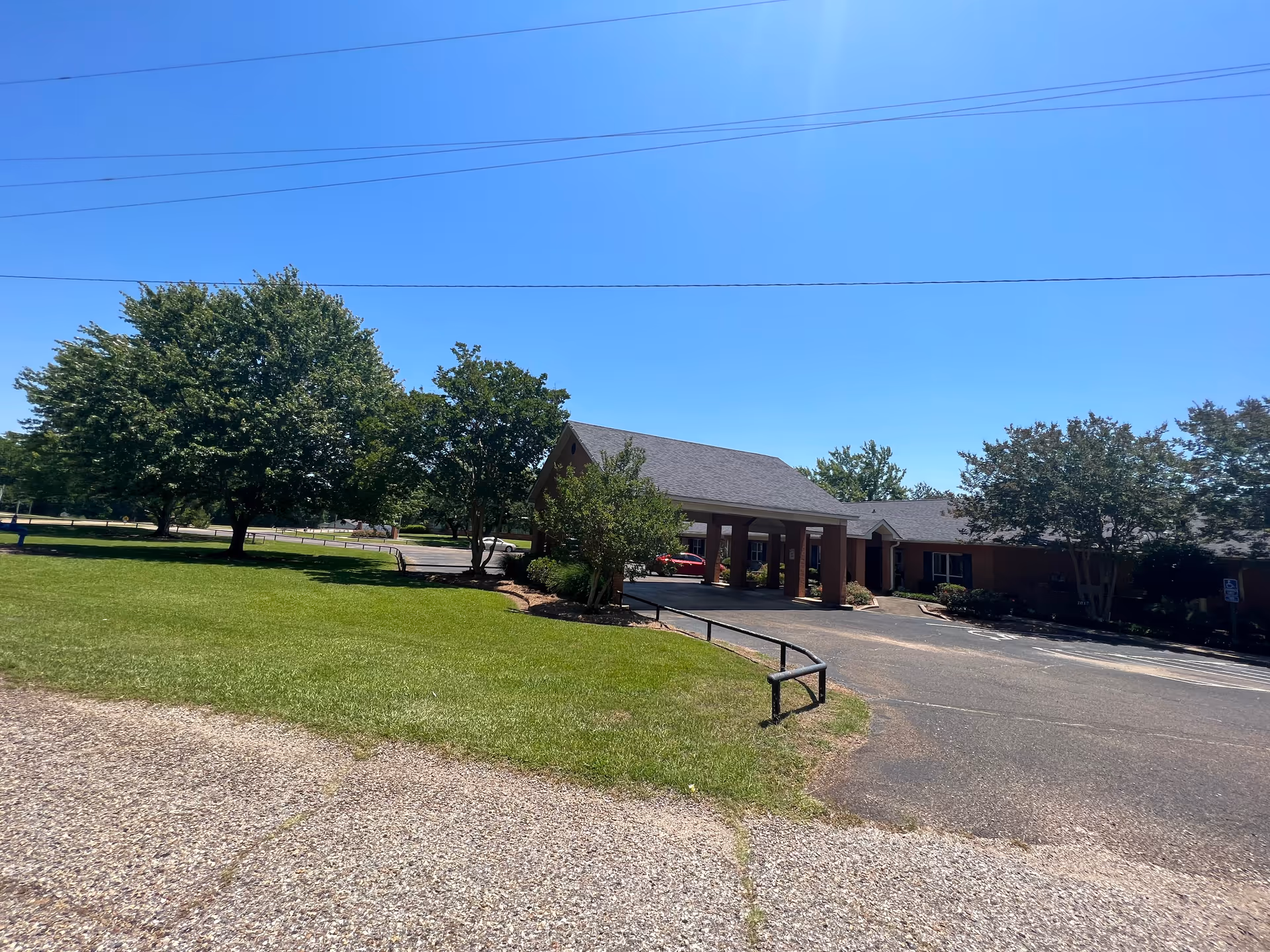 Exterior view of Leslie Lakes Retirement Center on a sunny day, showing a single-story building with a covered entrance, surrounded by green grass, trees, and a paved driveway.