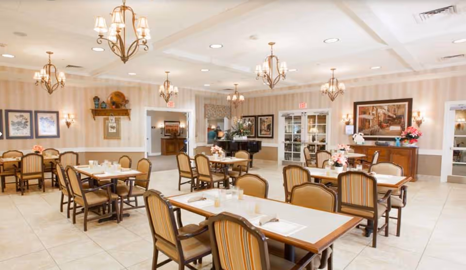A spacious dining room with multiple tables and chairs arranged neatly. The room features beige striped wallpaper, several framed pictures on the walls, and elegant chandeliers hanging from the ceiling. There are floral centerpieces on the tables and a piano in the background near a set of double glass doors.